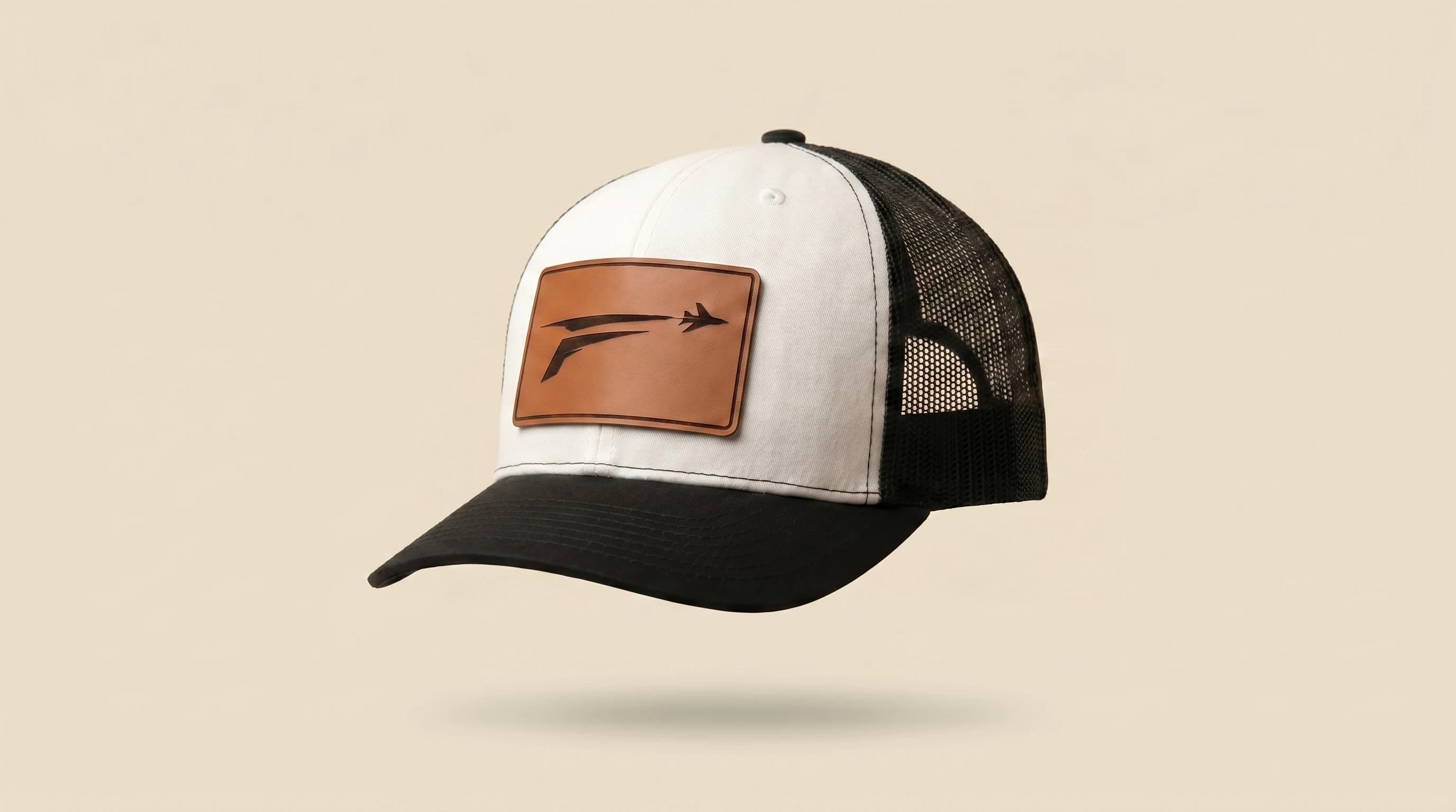The Icon trucker hat, three-quarter view. White panel, black bill, cognac leather patch with engraved F and plane mark.
